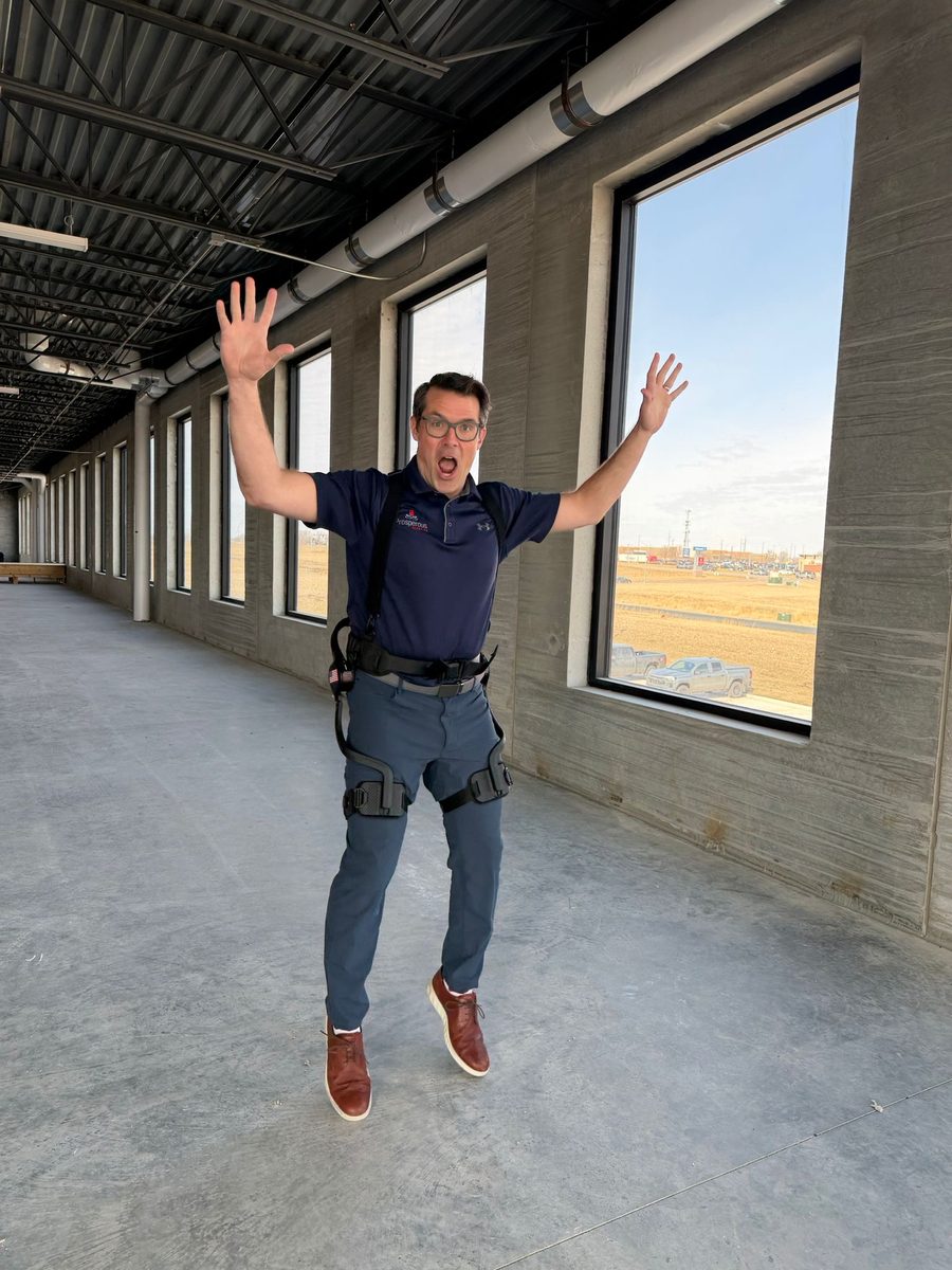Michael Bankowski jumping with both arms raised inside a long, unfinished concrete floor with exposed ceiling ducts and tall windows looking out onto open prairie. He is wearing a hip exoskeleton over his clothes.