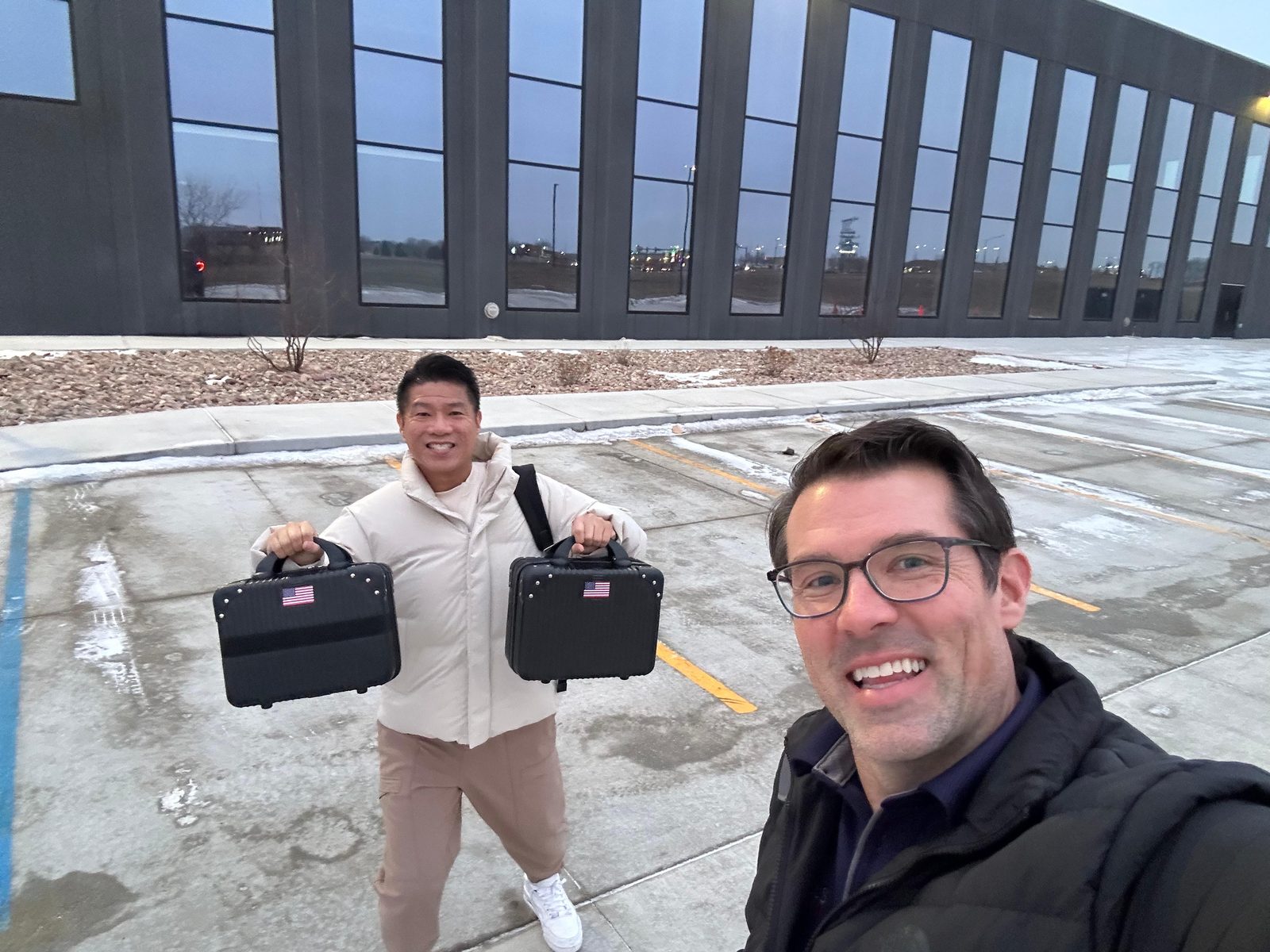 Zen Koh standing in a snowy parking lot outside the Discovery District building at dusk, holding up two black hard sided device cases, each marked with a small United States flag.