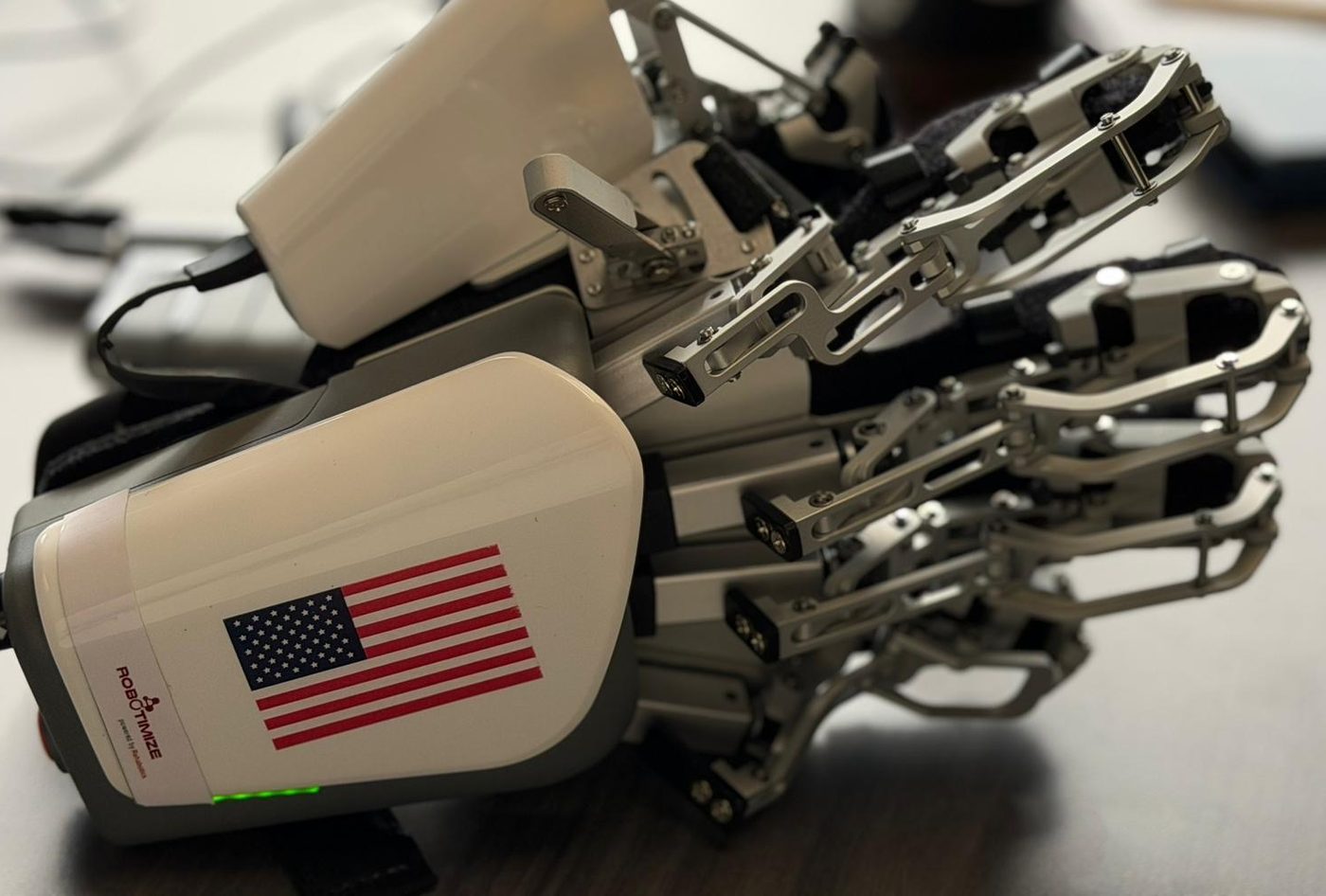 The HandVivante hand therapy device photographed on a desk, powered on with its indicator lit. A jointed aluminium linkage runs along each finger from a white housing carrying an American flag decal and the Robotimize maker mark.