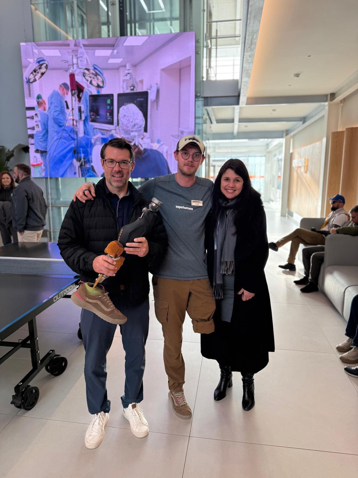 Michael Bankowski standing in the atrium of the Superhumans Center in Lviv with two other people. He is holding a working prosthetic leg that still has a sneaker on the foot and a small teddy bear tied to it. Beside him is Eddie, wearing a Superhumans shirt and a staff badge.