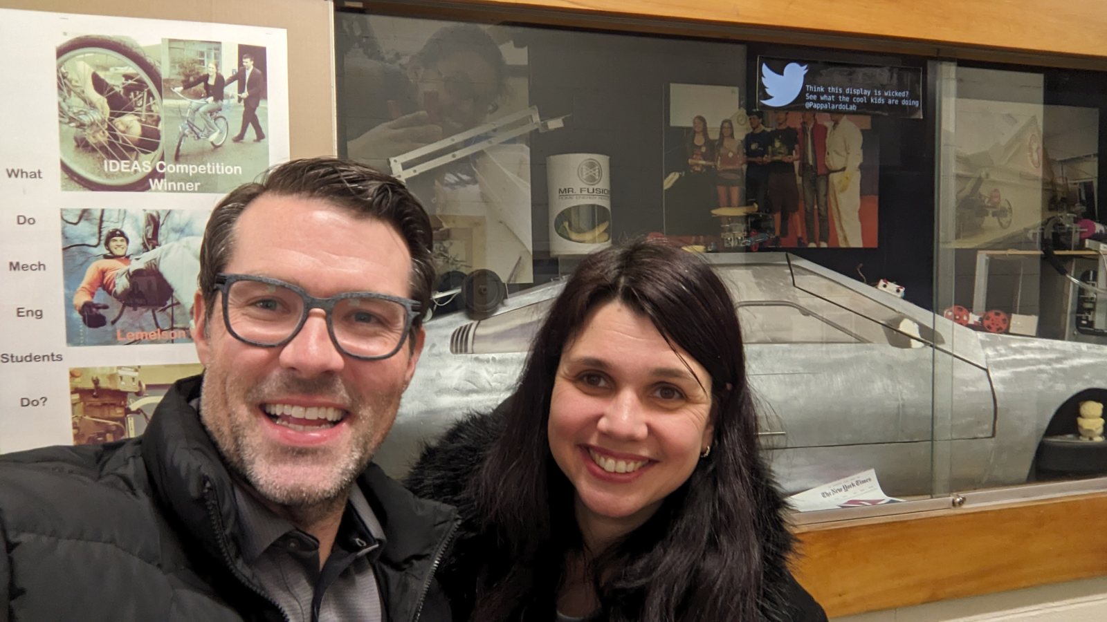 Michael Bankowski and Maja Rudinac standing in a corridor at the MIT Newman Lab, in front of a glass display case holding a partly built, bare metal DeLorean and a Mr. Fusion canister.