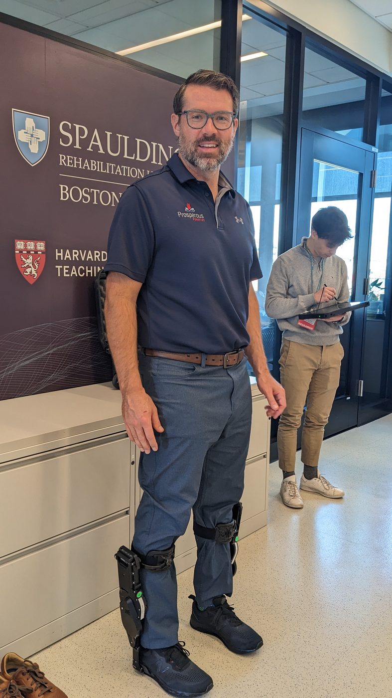 Michael Bankowski standing in front of a Spaulding Rehabilitation Hospital sign in Boston, wearing powered ankle modules strapped over his trousers and shoes. A researcher works at a tablet behind him.